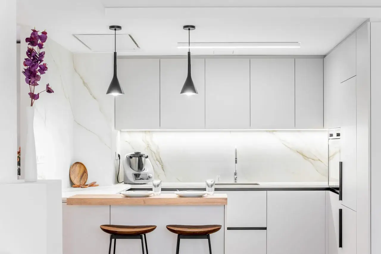 Moderna cocina pequeña con encimeras de mármol, gabinetes blancos y una barra de desayuno de madera.