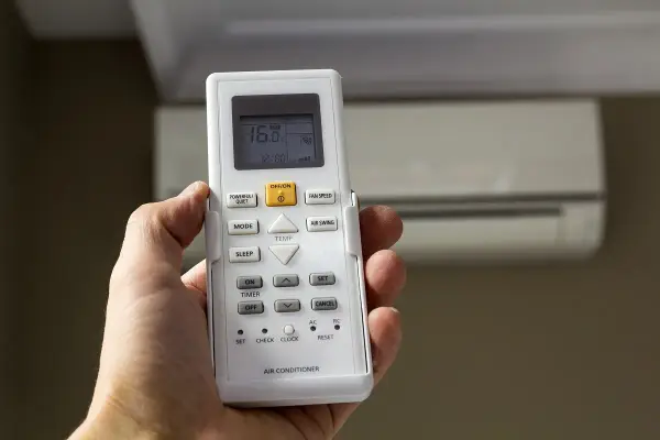 Mano sujetando mando para configurar mando universal aire acondicionado sin código. Aire acondicionado al fondo.