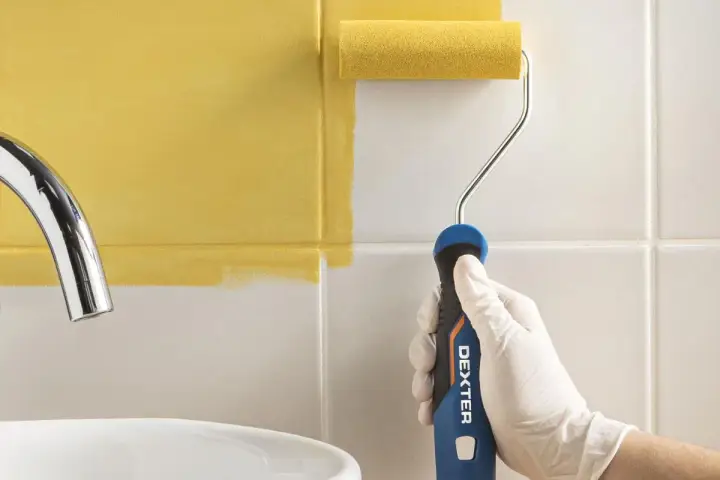 Mano con guante blanco pintando azulejos de cocina de blanco a amarillo con rodillo.