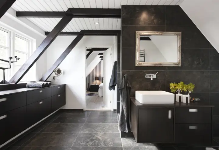 Baño moderno con vigas oscuras y suelo gris. El lavabo sobre encimera y los muebles oscuros contrastan con las paredes de baldosas oscuras. Se ve un dormitorio al fondo.